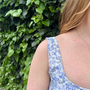 May include: A close-up of a woman's shoulder and chest wearing a white and blue floral print sleeveless top. The top has a scoop neck and the straps are made of the same fabric as the top.
