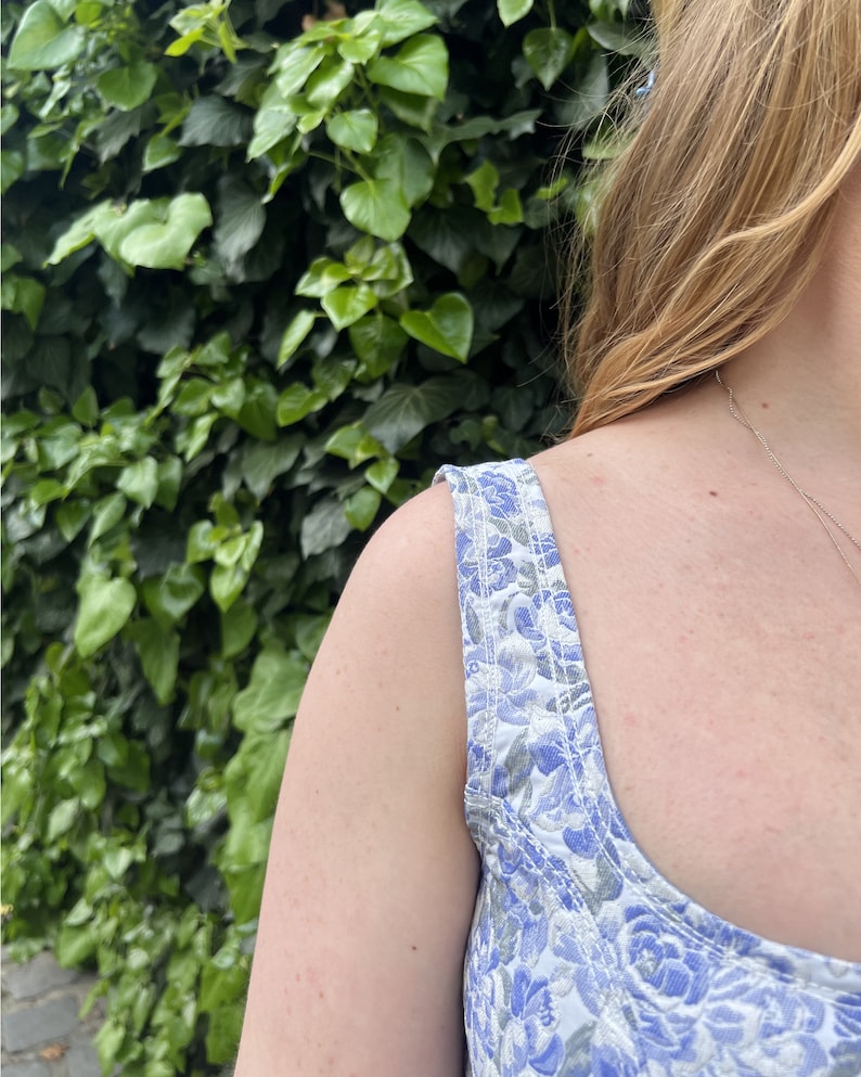 May include: A close-up of a woman's shoulder and chest wearing a white and blue floral print sleeveless top. The top has a scoop neck and the straps are made of the same fabric as the top.