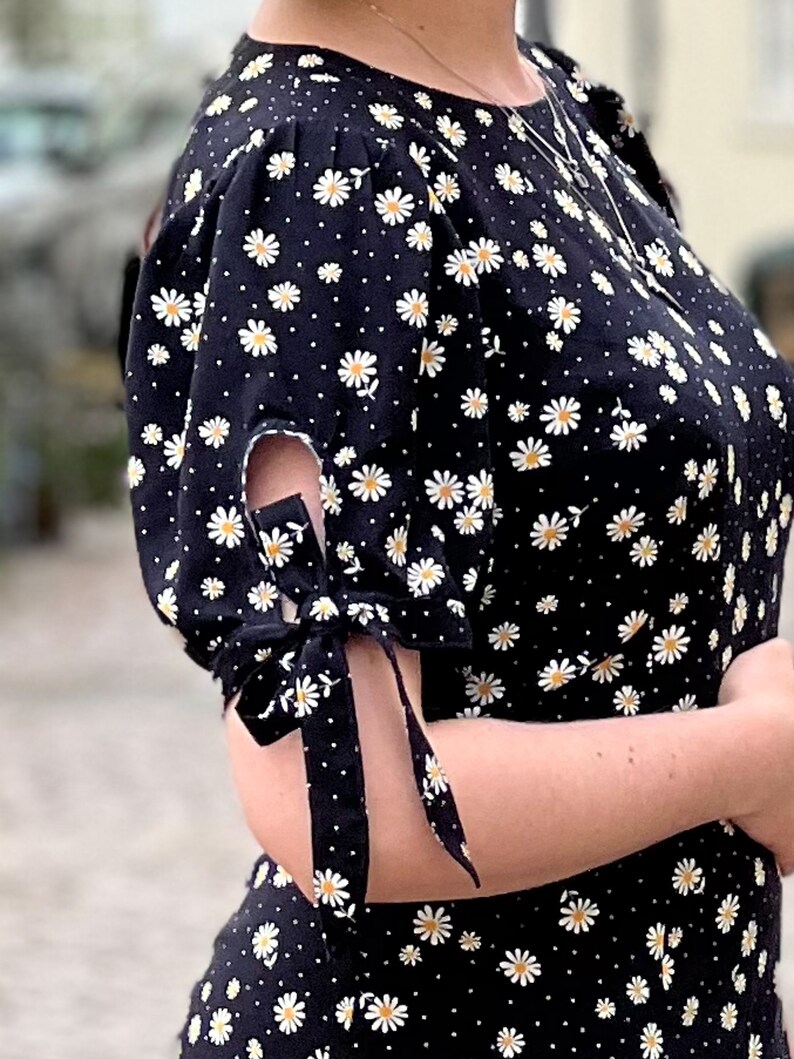 Puede incluir: Vestido negro con estampado de margaritas blancas y mangas atadas. El vestido tiene un escote redondo y una silueta ajustada.