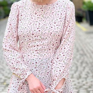 Puede incluir: Un vestido rosa de manga larga con estampado floral. El vestido tiene un escote redondo y pu&ntilde;os con cordones.