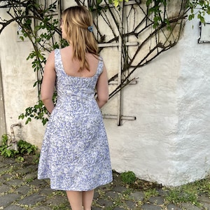 May include: A woman wearing a white dress with a blue floral pattern. The dress has a square neckline and a fitted bodice. The skirt is full and flows to the knees. The woman is standing in front of a white wall with a vine growing on it.