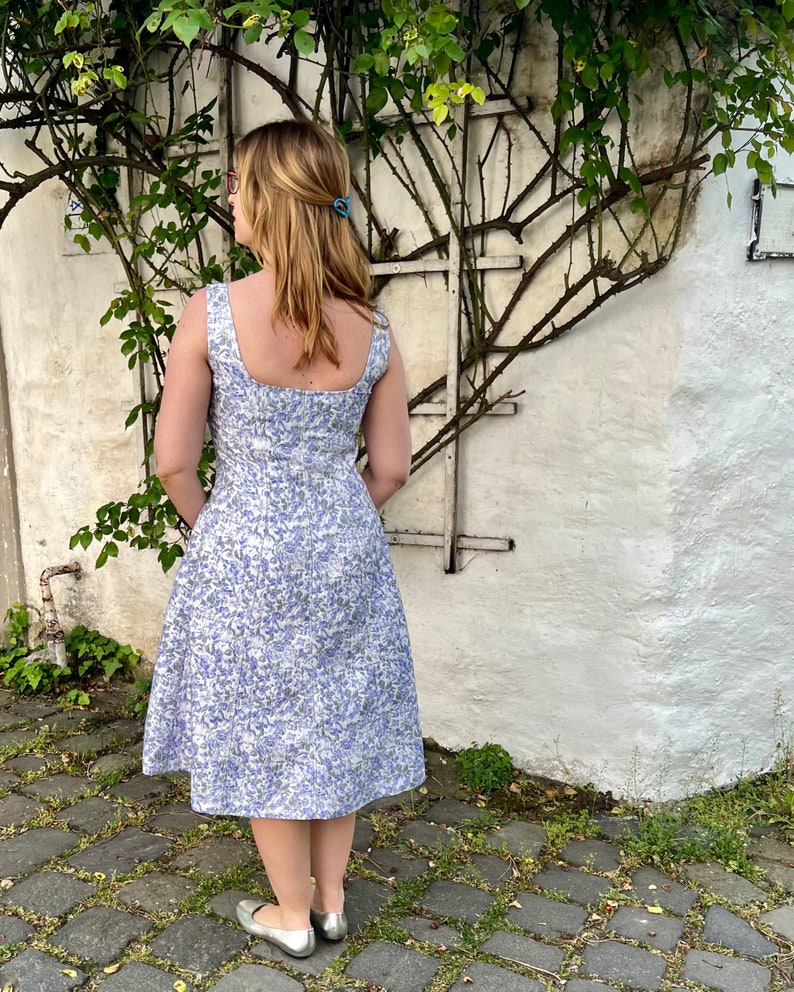May include: A woman wearing a white dress with a blue floral pattern. The dress has a square neckline and a fitted bodice. The skirt is full and flows to the knees. The woman is standing in front of a white wall with a vine growing on it.
