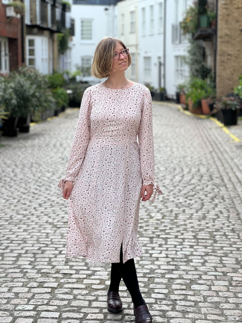 Puede incluir: Una mujer lleva un vestido midi de flores rosas con mangas largas y una peque&ntilde;a abertura en la falda. Lleva medias negras y mocasines marrones. El vestido tiene un corpi&ntilde;o ajustado y una falda fluida.