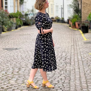 Puede incluir: Una mujer con un vestido midi negro con estampado floral blanco y sandalias amarillas camina por una calle adoquinada.