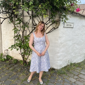May include: A woman wearing a light blue floral dress and silver flats stands in front of a white building with a sign that reads "Kieserica".
