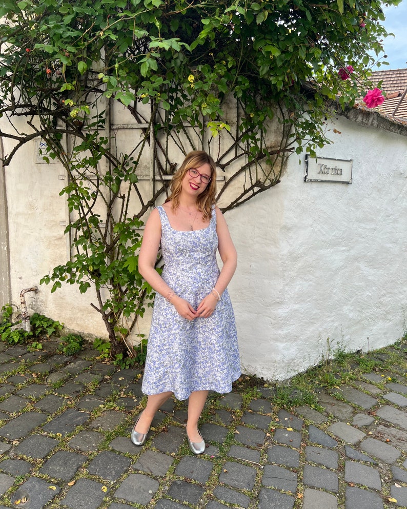 May include: A woman wearing a light blue floral dress and silver flats stands in front of a white building with a sign that reads "Kieserica".
