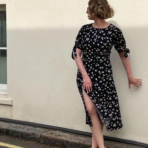 Puede incluir: Una mujer con un vestido midi negro con estampado floral blanco y sandalias de tac&oacute;n amarillo. El vestido tiene una abertura lateral y las mangas est&aacute;n atadas en las mu&ntilde;ecas.