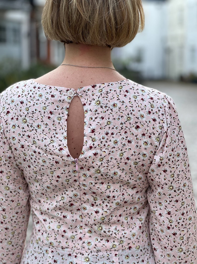 Puede incluir: Un vestido rosa floral con un escote en la espalda en forma de ojo de cerradura y una cremallera. El vestido tiene un peque&ntilde;o estampado floral con flores blancas y hojas verdes sobre un fondo rosa.