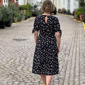 Puede incluir: Una mujer lleva un vestido midi negro con estampado floral blanco. El vestido tiene un escote en la espalda y mangas atadas. Est&aacute; caminando por una calle adoquinada.