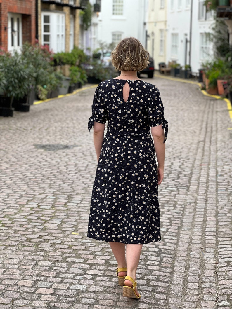 Puede incluir: Una mujer lleva un vestido midi negro con estampado floral blanco. El vestido tiene un escote en la espalda y mangas atadas. Est&aacute; caminando por una calle adoquinada.