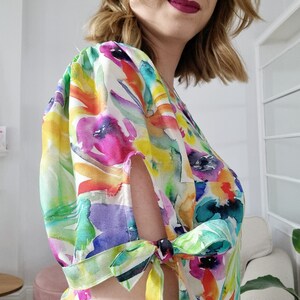 Puede incluir: Un vestido blanco con estampado floral colorido y mangas abullonadas. Las mangas tienen un detalle de lazo en la mu&ntilde;eca.
