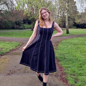 May include: A woman wearing a black midi dress with white stitching detail and black ankle boots. She is standing on a path in a park.