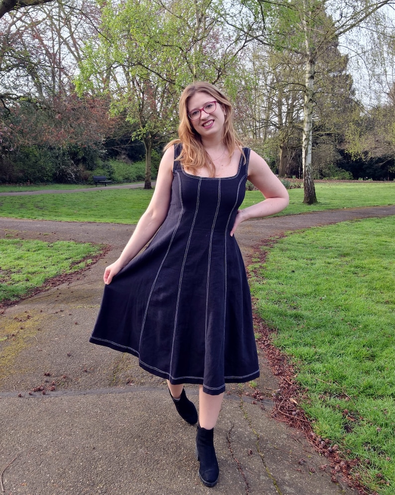 May include: A woman wearing a black midi dress with white stitching detail and black ankle boots. She is standing on a path in a park.