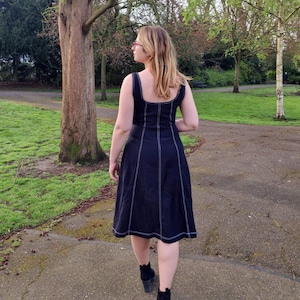 May include: A woman wearing a black midi dress with white stitching detail and black ankle boots. She is standing in a park with green grass and trees in the background.