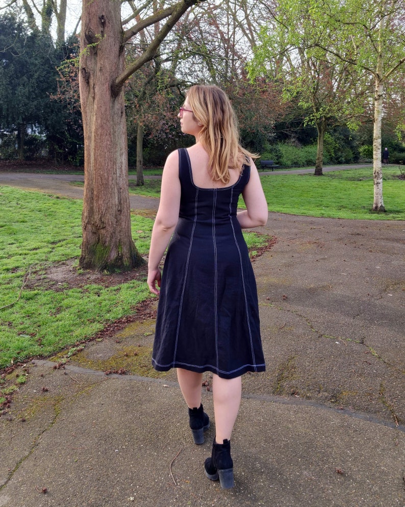 May include: A woman wearing a black midi dress with white stitching detail and black ankle boots. She is standing in a park with green grass and trees in the background.