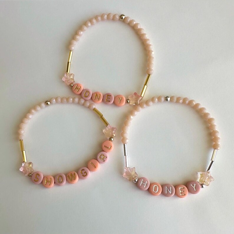 May include: Three beaded bracelets with the words "HONEY" and "SHOWGIRL" spelled out in pink and peach letter beads. The bracelets have gold and silver accents, and star-shaped charms. The beads are a light pink color.