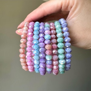 May include: A stack of pastel-colored bracelets made of faceted glass beads. The bracelets are arranged in a rainbow order, starting with pink and ending with purple.