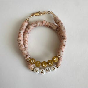 Peut inclure: Deux bracelets superposés avec des perles en forme de disque roses et beiges. Le bracelet supérieur comporte des perles dorées épelant "RILEY", et le bracelet inférieur comporte des perles blanches épelant "MAMA". Les bracelets sont reliés par un fermoir doré.