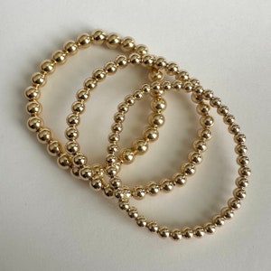 May include: Three gold beaded bracelets stacked on top of each other. The bracelets are made of small, round beads.