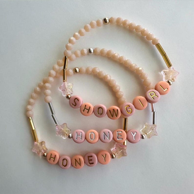 May include: Three beaded bracelets with the words "SHOWGIRL" and "HONEY" spelled out in colorful letter beads. The bracelets feature peach, pink, and gold accents, with star-shaped charms. The beads are a light peach color.