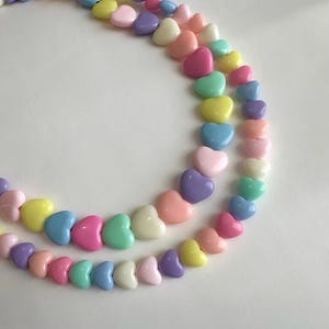 May include: Two strands of heart-shaped beads in a variety of pastel colors, including pink, yellow, blue, green, and purple. The beads are arranged in a curved pattern against a white background.