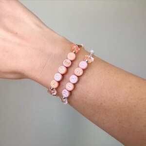 May include: Two beaded bracelets spelling "HONEY" in pastel pink, peach, and yellow letter beads. The bracelets have clear crystal accents and are worn on a wrist. The background is a solid light gray.