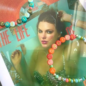 May include: Framed artwork featuring a portrait of a woman with beaded necklaces spelling out "THE LIFE OF SHOWGIRL". The background includes a vinyl record and text. The necklaces have colorful beads and lettered discs.