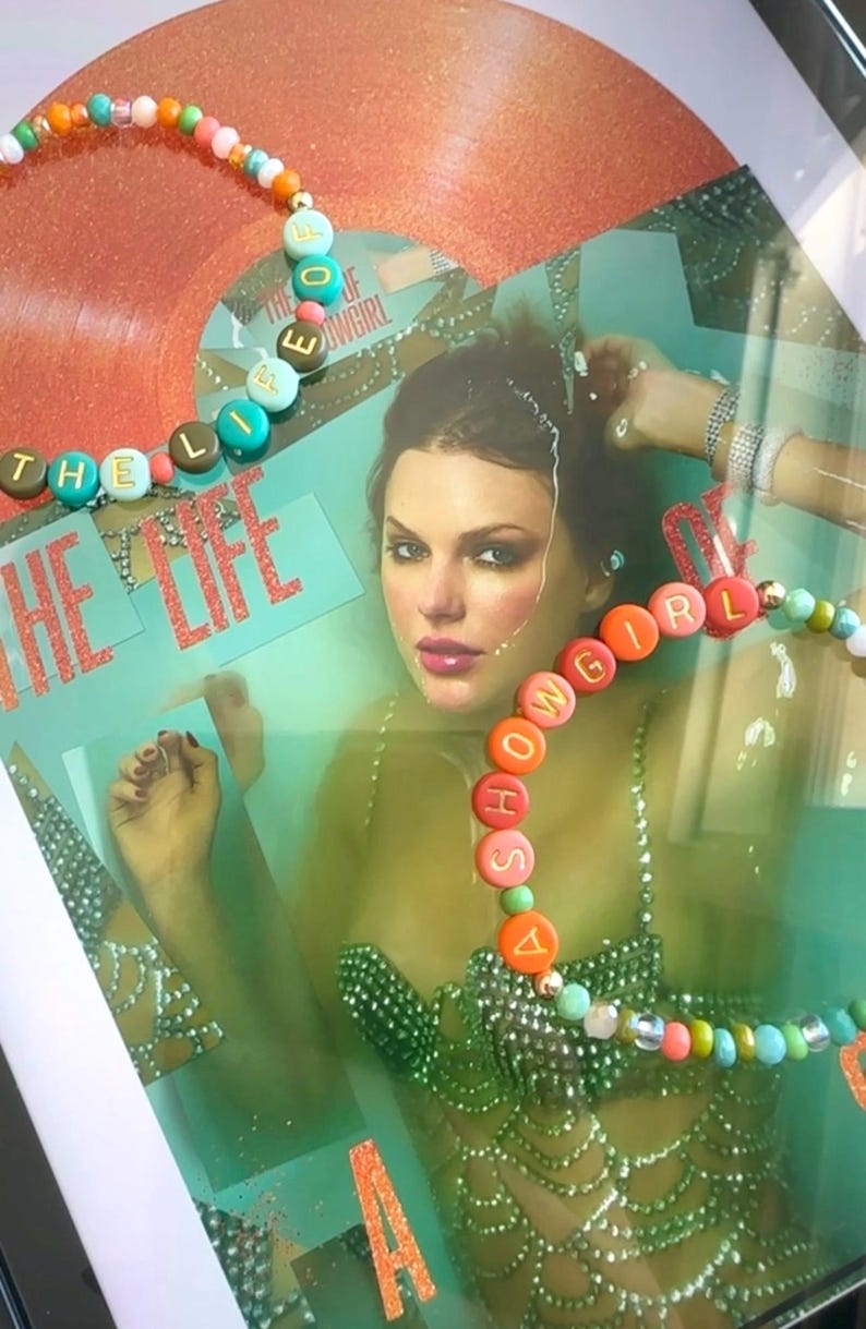 May include: Framed artwork featuring a portrait of a woman with beaded necklaces spelling out "THE LIFE OF SHOWGIRL". The background includes a vinyl record and text. The necklaces have colorful beads and lettered discs.