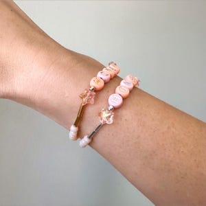 May include: Two beaded bracelets with the word "HOPE" spelled out in white letter beads on pink beads. Each bracelet has a star-shaped charm and a gold or silver-colored metal rod.