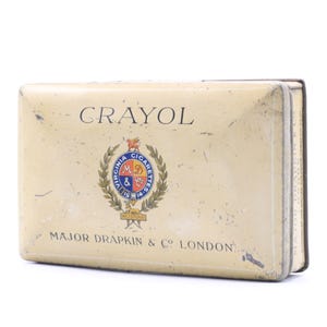 May include: A vintage tin box for Crayol Virginia Cigarettes. The box is cream colored with gold lettering and a crest featuring a blue shield with the letters "M & D" and the words "Virginia Cigarettes" in a circle. The text on the box reads "Crayol" and "Major Draffin & Co. London."