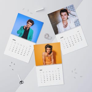 Op de afbeelding: Een wandkalender voor 2027 met drie pagina's met foto's van een persoon. De april-pagina toont een persoon in een groen jasje, de mei-pagina in een oranje tuinbroek en de juni-pagina in een wit jasje. Elke pagina bevat een maandoverzicht.