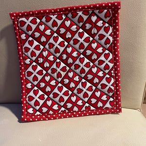 May include: Red and white quilted pot holder with a heart pattern and white polka dots around the border.