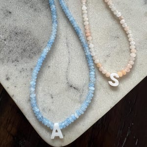 Peut inclure: Deux colliers de perles. L'un est bleu clair avec la lettre blanche "A", l'autre est rose avec la lettre blanche "S". Les colliers sont sur une surface blanche et grise.