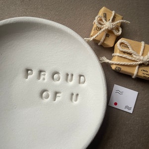 May include: White ceramic dish with the words "PROUD OF U" embossed on the surface. Two small, brown, wrapped packages are next to the dish.
