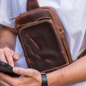 Könnte beinhalten: Eine braune Leder-Umhängetasche mit Reißverschluss und einem Used-Look. Die Tasche wird von einer Person getragen, die ein Smartphone benutzt.