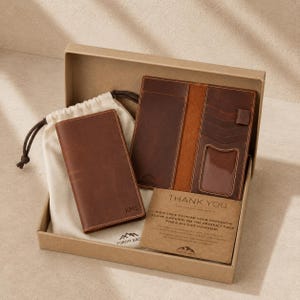 May include: A brown leather wallet set in a cardboard box, including a wallet, a passport holder, and a thank you card. The passport holder has a clear ID window. The wallet is embossed with the initials "KMS".