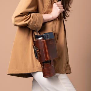Op de afbeelding: Een bruine leren schoudertas met een telefoon- en een bekerhouder. De tas heeft een band en een gespsluiting. De tas wordt gedragen door een persoon met bruin haar.