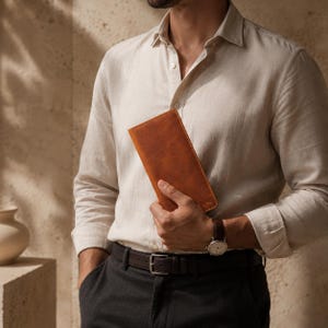 Pode incluir: Uma carteira de couro marrom é segurada na mão. A carteira é retangular e parece ser feita de couro de alta qualidade. A pessoa está vestindo uma camisa clara abotoada e um relógio com uma pulseira de couro marrom.