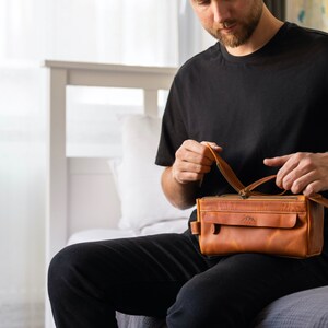 A male traveler sitting on a bed, holding the rugged leather toiletry case for his journey.