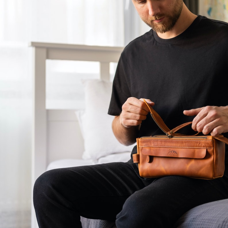 A male traveler sitting on a bed, holding the rugged leather toiletry case for his journey.
