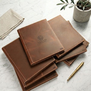 May include: A stack of brown leather journals with stitched edges and the "Crafted Leatherworks" logo. Each journal has initials embossed on the bottom right corner. A gold pen rests on the marble surface next to the journals.