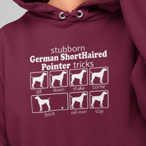 May include: A maroon hoodie featuring a white graphic of a German Shorthaired Pointer dog and the text "stubborn German Shorthaired Pointer tricks". The graphic depicts the dog in various poses with the words "sit", "down", "shake", "come", "fetch", "roll-over", and "stay" beneath each pose.