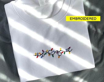 May include: White crewneck sweatshirt with an embroidered string of colorful Christmas lights. The word "EMBROIDERED" is in a yellow rectangle. The sweatshirt is folded on a gray surface.