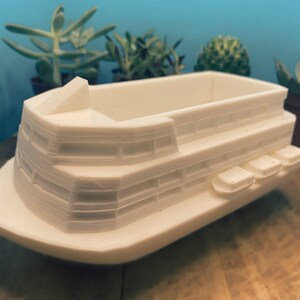 May include: White ceramic planter shaped like a boat with multiple decks. The planter has a rectangular opening for planting.