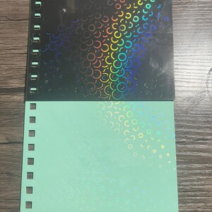 Puede incluir: Un cuaderno con espiral con una cubierta negra con un dise&ntilde;o hologr&aacute;fico de c&iacute;rculos superpuestos. La mitad inferior del cuaderno es de color verde claro con un patr&oacute;n hologr&aacute;fico similar. El cuaderno tiene un borde perforado.