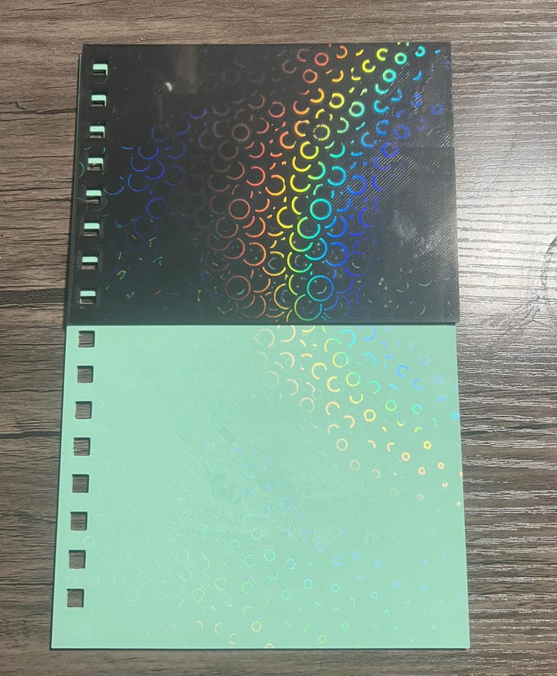 Puede incluir: Un cuaderno con espiral con una cubierta negra con un dise&ntilde;o hologr&aacute;fico de c&iacute;rculos superpuestos. La mitad inferior del cuaderno es de color verde claro con un patr&oacute;n hologr&aacute;fico similar. El cuaderno tiene un borde perforado.