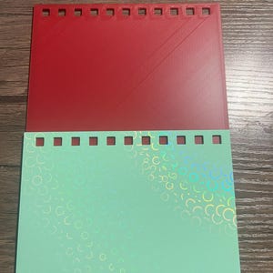 Puede incluir: Un juego de papeler&iacute;a de dos piezas con una parte superior roja y una inferior verde menta. La pieza roja tiene una superficie texturizada y una fila de recortes cuadrados a lo largo del borde superior. La pieza verde menta tiene un patr&oacute;n hologr&aacute;fico de c&iacute;rculos superpuestos.
