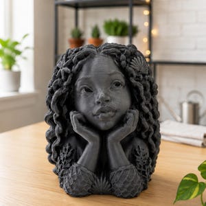 Peut inclure: Une jardinière sculpturale gris foncé en forme de personne aux longs cheveux bouclés ornés de coquillages et d'étoiles de mer. La figure repose son menton sur ses mains, avec des manches texturées en filet et un coquillage à la base.