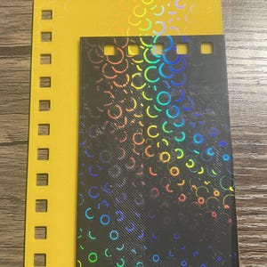 Puede incluir: Dos tarjetas rectangulares. Una es amarilla con recortes cuadrados a lo largo del borde izquierdo. La otra es negra con un patr&oacute;n de arco&iacute;ris hologr&aacute;fico de c&iacute;rculos y formas de C. Las tarjetas est&aacute;n superpuestas.