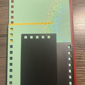 Puede incluir: Un surtido de hojas de papel de colores, incluyendo verde, negro y rojo, con bordes perforados para encuadernar. La hoja verde tiene un patr&oacute;n hologr&aacute;fico iridiscente. Las hojas est&aacute;n dispuestas sobre una superficie de madera.